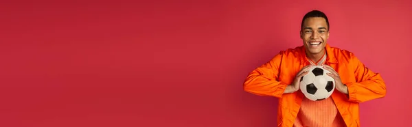 Щасливий афроамериканський чоловік в помаранчевій сорочці тримає футбольний м'яч на червоному, банер, копіювальний простір — стокове фото