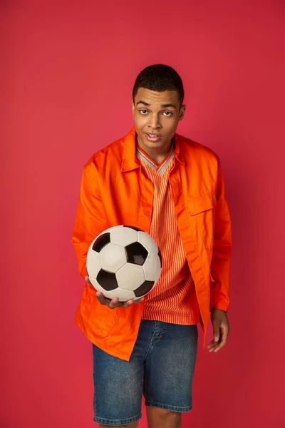 Улыбающийся и скептически настроенный африканский американец в оранжевой рубашке показывает футбольный мяч на красном фоне — стоковое фото