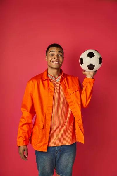 Fröhlicher afrikanisch-amerikanischer Mann in orangefarbenem Hemd, Fußballball in der Hand und lächelnd in die Kamera auf rot — Stockfoto