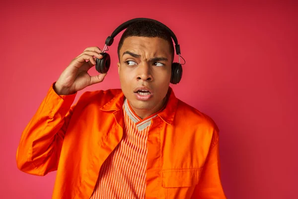 Surprised african american guy in orange shirt listening music in wireless headphones on red — Stock Photo