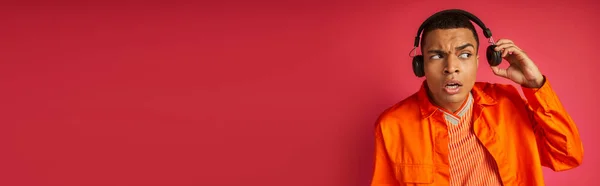 Überraschte afrikanisch-amerikanische Mann in drahtlosen Kopfhörern und orangefarbenem Hemd auf rot, Banner, Kopierraum — Stockfoto