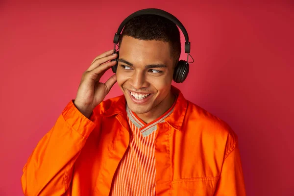 Sonriente hombre afroamericano en camisa naranja y auriculares inalámbricos escuchando música en rojo - foto de stock