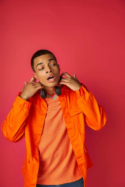 Hombre afroamericano sorprendido en camisa naranja, con auriculares inalámbricos mirando hacia otro lado en rojo - foto de stock