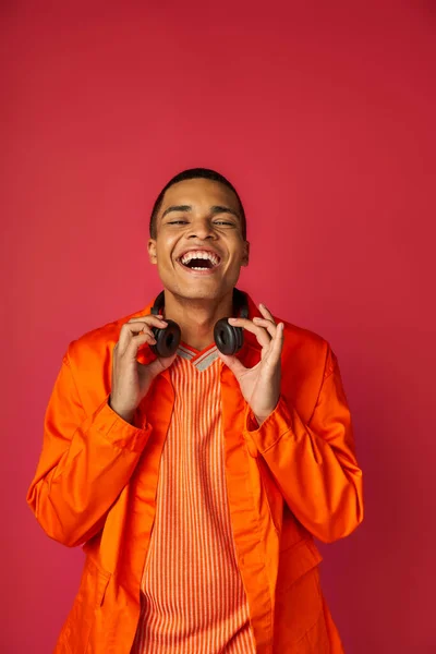 Stylish african american man in orange shirt, with wireless headphones laughing on red — Stock Photo