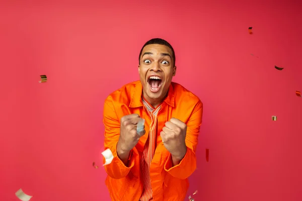 Overjoyed african american man in orange shirt shouting and showing win gesture near confetti on red — Stock Photo
