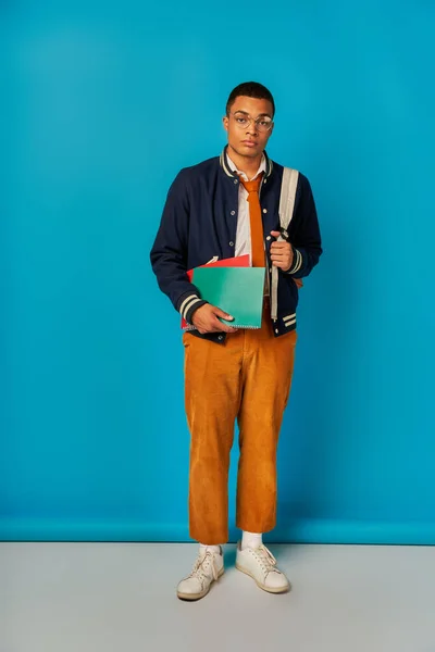 Étudiant afro-américain élégant avec sac à dos et cahiers regardant la caméra sur fond bleu — Photo de stock