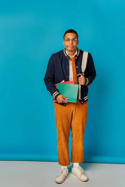 Annoiato africano americano studente con notebook e zaino sbuffare guance su sfondo blu — Foto stock