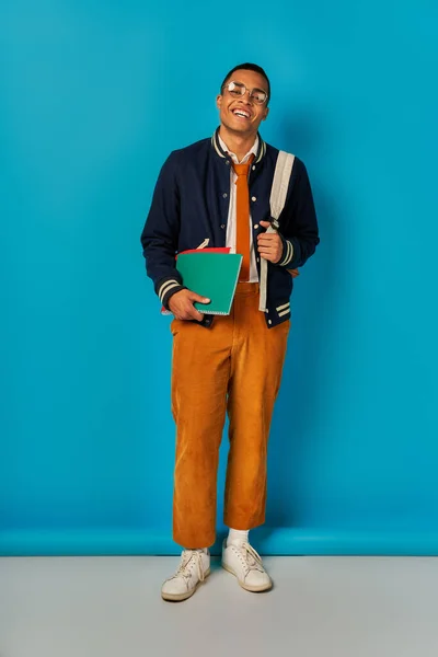 Cheerful african american student in jacket and orange pants standing with notebooks on blue — Stock Photo