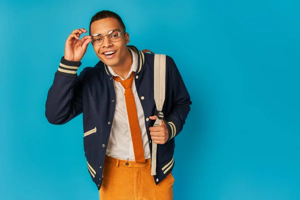 Stylish african american student in eyeglasses smiling at camera on blue, notebooks, backpack — Stock Photo