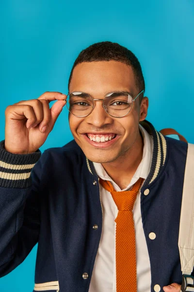 Cheerful and trendy african american student in jacket, tie and eyeglasses smiling at camera on blue — Stock Photo