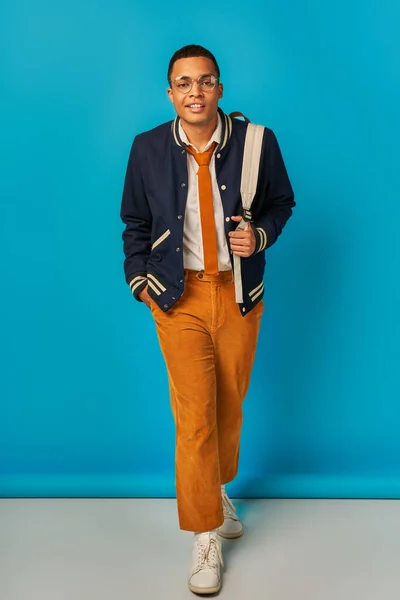 Stylish african american student in jacket and orange pants walking with hand in pocket on blue — Stock Photo