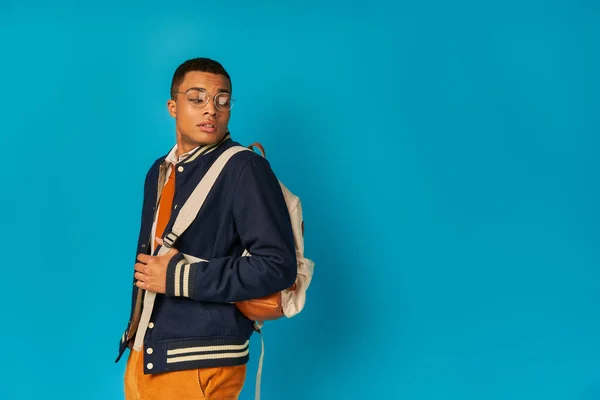Estudiante afroamericano de moda en gafas y chaqueta mirando la mochila sobre fondo azul - foto de stock