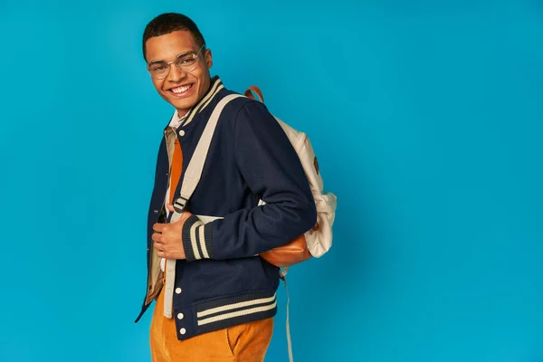 Carefree african american student in jacket and orange pant, with backpack looking at camera on blue — Stock Photo