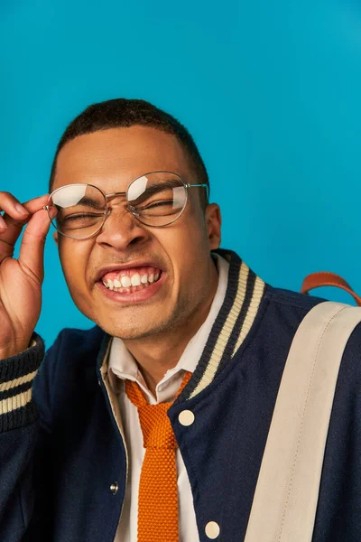 Funny african american student in jacket adjusting eyeglasses and grimacing on blue background — Stock Photo