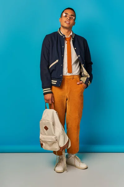 Уверенный в себе африканский студент в модной повседневной одежде с рюкзаком и рукой в кармане на голубом — стоковое фото