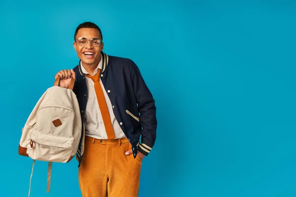 Excited african american student in trendy jacket and orange pants holding backpack on blue — Stock Photo
