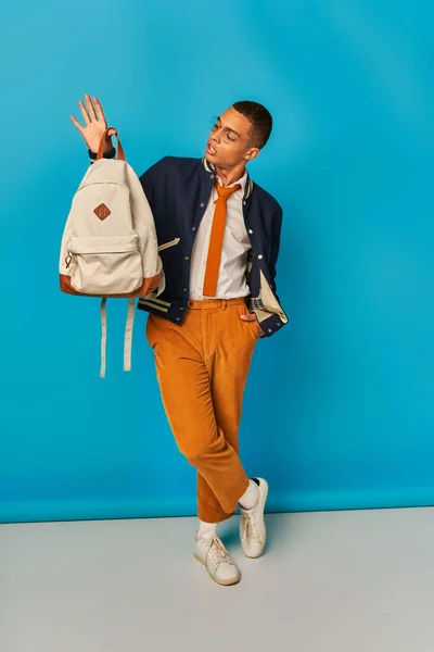 African american student in orange pants standing with hand in pocket and backpack on blue — Stock Photo