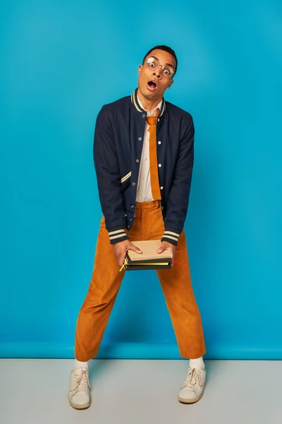 Schockierter afrikanisch-amerikanischer Student mit offenem Mund, Notizbücher auf blauem Grund, Jacke, orangefarbene Hose — Stockfoto