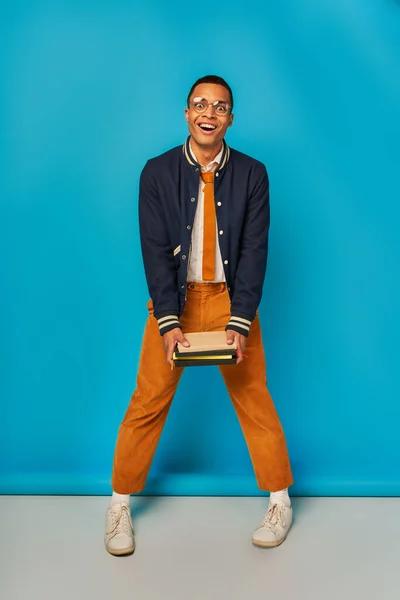 Surprised african american student in trendy jacket and orange pants standing with notebooks on blue — Stock Photo