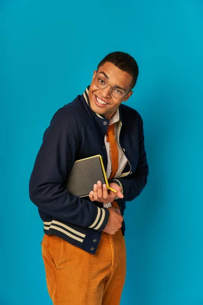 Cheerful and tricky african american student with notebooks looking at camera on blue, trendy jacket — Stock Photo