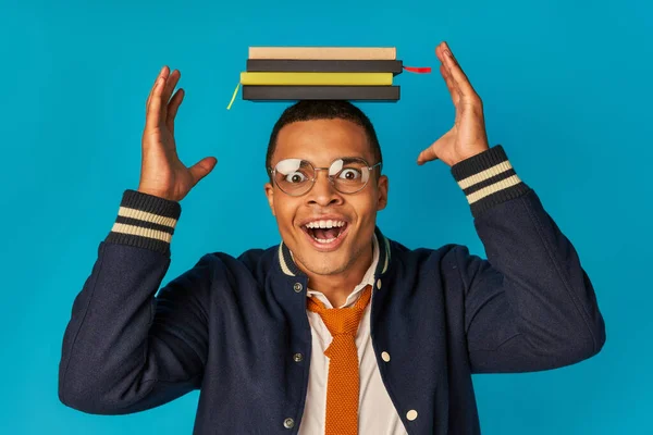 Amazed african american student posing with notebooks on head on blue, eyeglasses, stylish jacket — Stock Photo