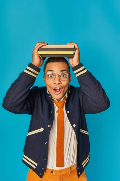 Étudiant afro-américain étonné dans une veste à la mode et des lunettes avec des cahiers sur la tête sur bleu — Photo de stock