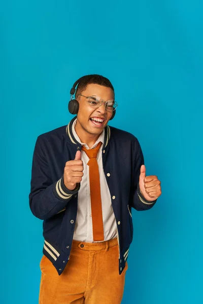 Estudiante afroamericano emocionado en auriculares inalámbricos y chaqueta de moda que muestra los pulgares hacia arriba en azul - foto de stock