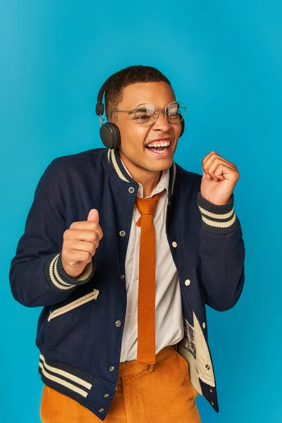 Overjoyed african american student in trendy jacket listening music in wireless headphones on blue — Stock Photo