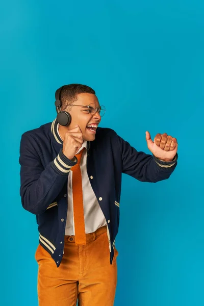 Energized, stylish african american student in wireless headphones listening music on blue — Stock Photo