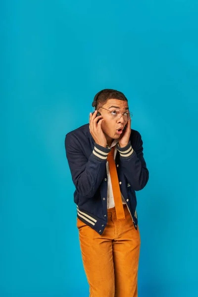 Asombrado estudiante afroamericano en chaqueta escuchando música en auriculares inalámbricos en azul - foto de stock