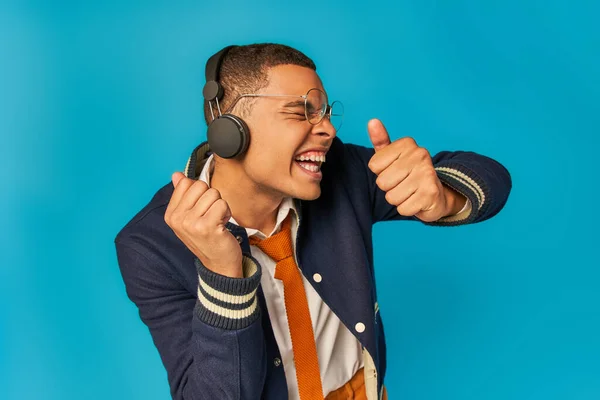 Étudiant afro-américain expressif écouter de la musique dans les écouteurs et montrant pouces vers le haut sur le bleu — Photo de stock