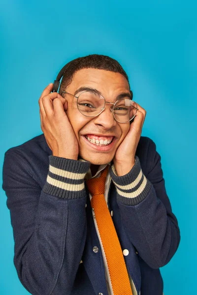 Alegre afroamericano estudiante en gafas, chaqueta y auriculares mirando a la cámara en azul - foto de stock