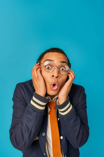 Choqué étudiant afro-américain en lunettes et écouteurs regardant la caméra sur bleu, bouche ouverte — Photo de stock
