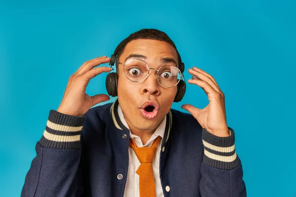 Impressed african american student in eyeglasses and headphones, with open mouth on blue — Stock Photo
