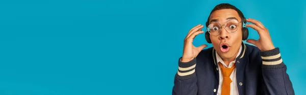 Shocked african american student in trendy jacket, eyeglasses and headphones on blue, banner — Stock Photo