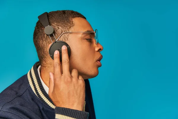 Profile of african american student in jacket and eyeglasses touching wireless headphones on blue — Stock Photo