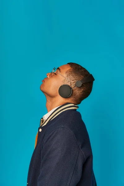Vista lateral del estudiante afroamericano de moda en chaqueta escuchando música en auriculares en azul - foto de stock