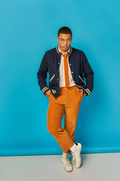 Confident african american student in jacket and orange pants posing with hands in pockets on blue — Stock Photo