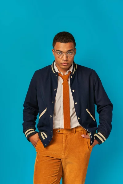 Modern, trendy african american student in jacket, orange pants, with hands in pockets on blue — Stock Photo