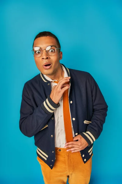 Shocked african american student in eyeglasses adjusting tie and looking at camera on blue — Stock Photo