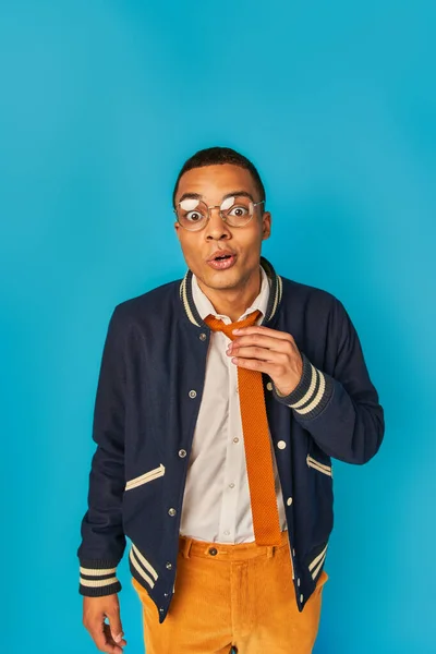 Surpreso e moderno estudante afro-americano em jaqueta tocando gravata e olhando para a câmera em azul — Fotografia de Stock