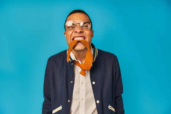 African american student in stylish jacket and eyeglasses having fun and biting tie on blue — Stock Photo