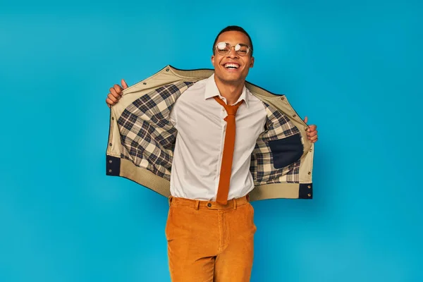 Étudiant afro-américain joyeux en veste d'ouverture de vêtements à la mode et souriant à la caméra sur bleu — Photo de stock