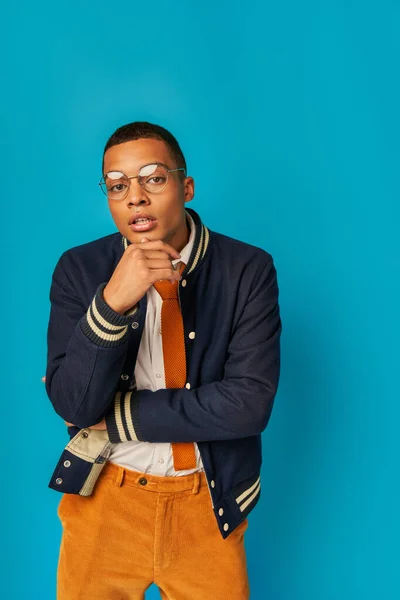 Pensativo africano americano estudiante en gafas y chaqueta de moda mirando a la cámara en azul - foto de stock