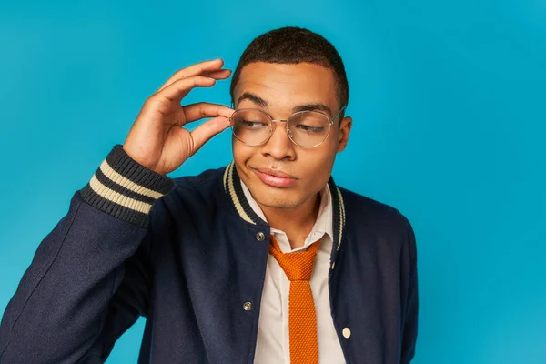 Estudiante afroamericano escéptico en chaqueta elegante ajustando gafas y mirando hacia otro lado en azul - foto de stock