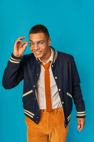 Carismático estudante afro-americano de jaqueta ajustando óculos e sorrindo para a câmera em azul — Fotografia de Stock