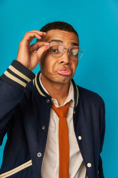Arrogante y elegante estudiante afroamericano ajustando gafas y mirando a la cámara en azul - foto de stock