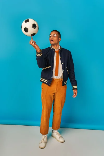 Studente afroamericano in giacca e pantaloni arancioni che gioca con pallone da calcio su blu — Foto stock