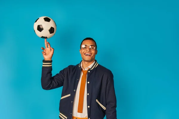 Alegre y elegante estudiante afroamericano jugando con la pelota de fútbol y mirando a la cámara en azul - foto de stock