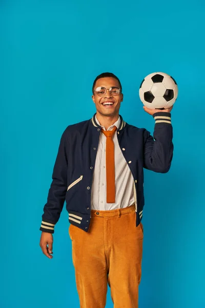 Estudiante afroamericano feliz en chaqueta y pantalones anaranjados jugando con pelota de fútbol en azul - foto de stock